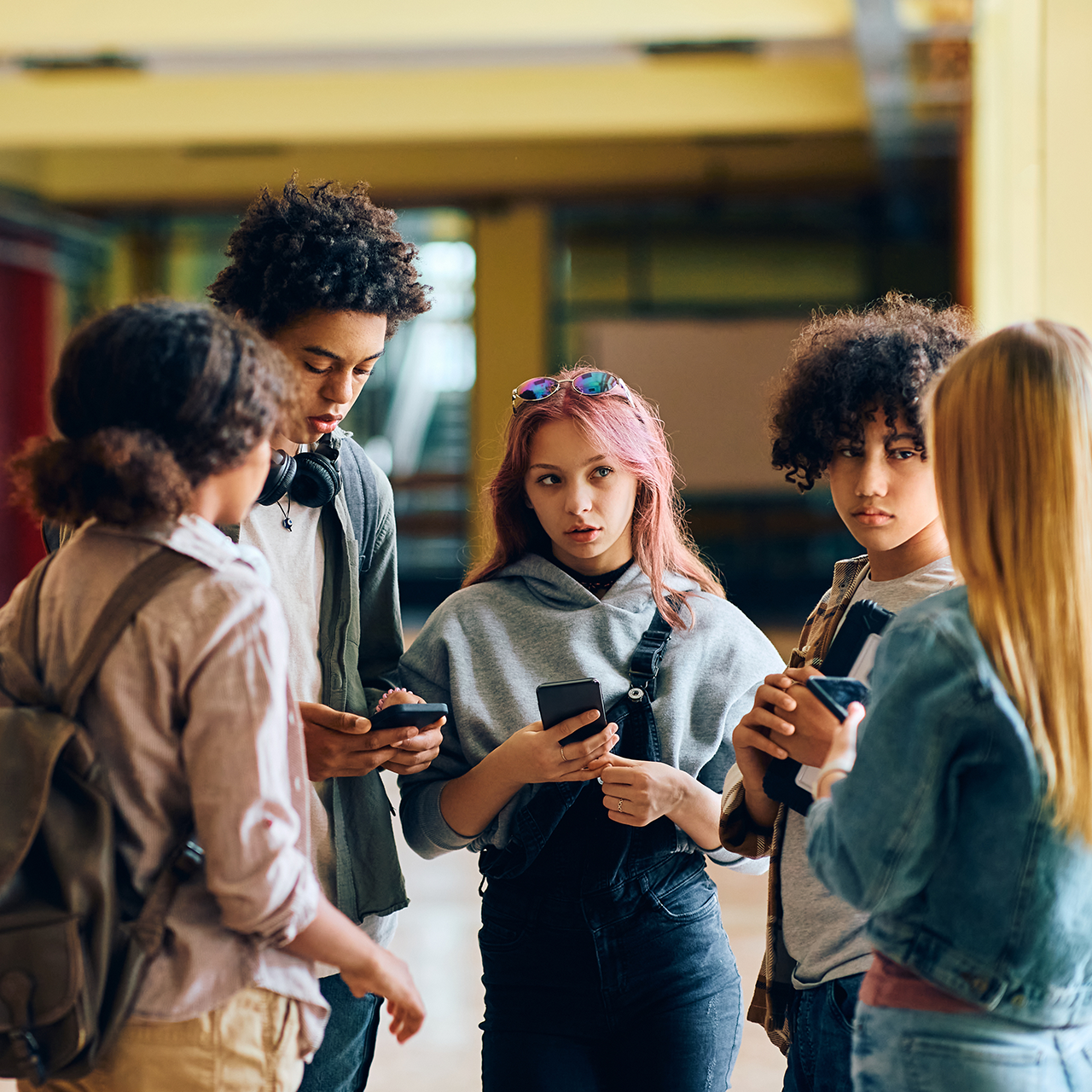 Fünf Jugendliche stehen in einem Schulflur zusammen, mehrere von ihnen halten Smartphones in der Hand und sprechen miteinander.